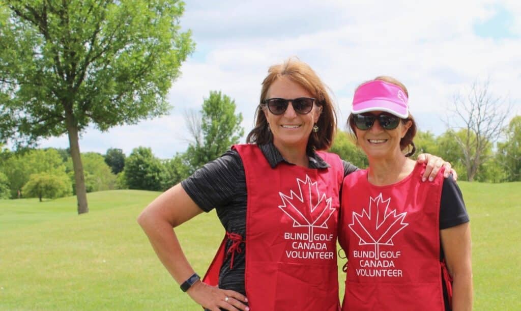 A beautiful sunny day, on a scenic golf hole with a tree in the background at The Greens at Renton. Two volunteers, with big smiles are wearing red bibs with a BGC maple leaf log and the words 'Blind Golf Canada Volunteer.'