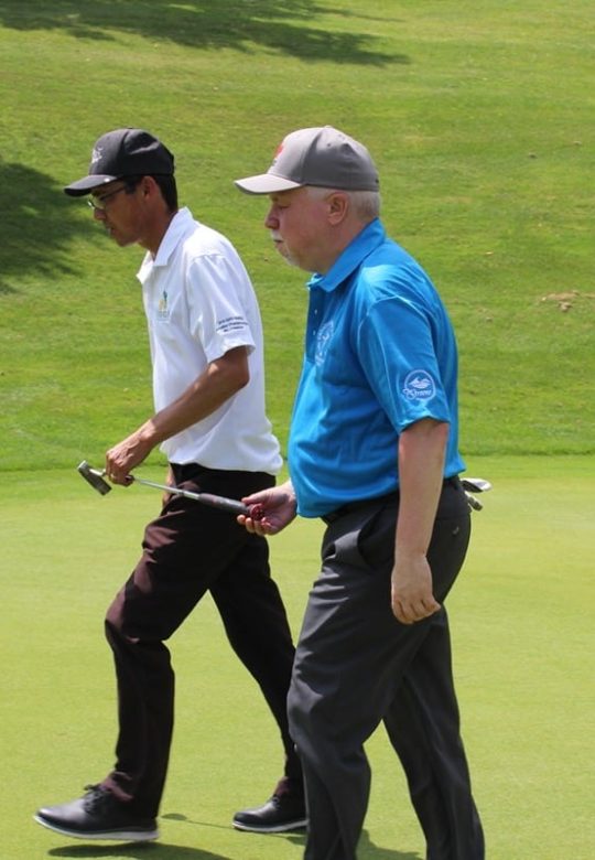 Two golfers are walking on a green approaching the hole. A blind coach in a white shirt, is guiding a B1 golfer using a putter. The putter acts as a stick that helps direct the blind golfer.